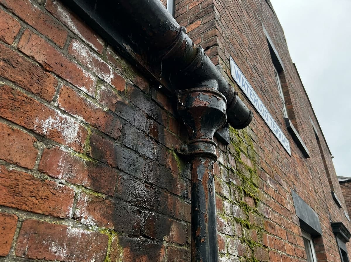 Dark damp patch spreading across exterior brickwork beneath a leaking gutter and downpipe